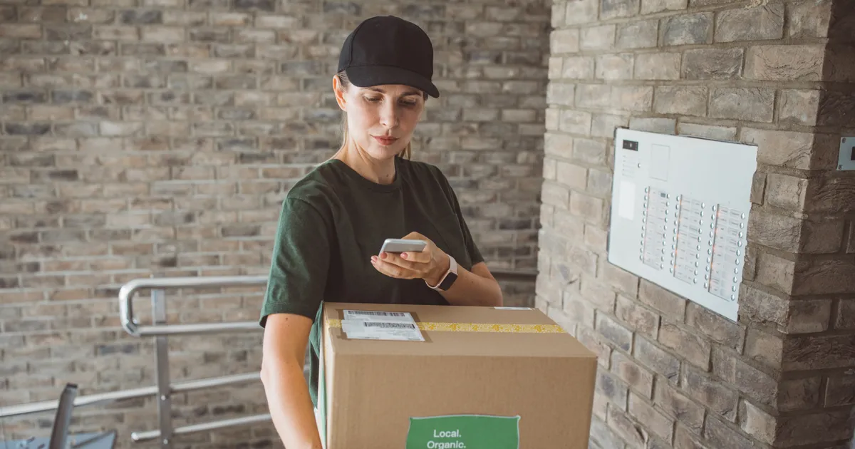 Delivery woman carrying box loaded with organic vegetables to the customer. Everything is freshly harvested from garden. She using mobile phone to check is she on right location